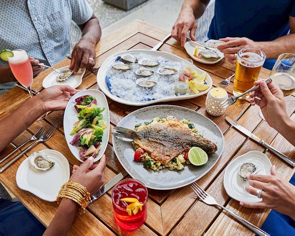 A group of friends enjoying a meal outdoors: oysters, a fish dish, salads, drinks, and shared plates around a wooden table.