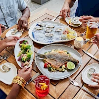 A group of friends enjoying a meal outdoors: oysters, a fish dish, salads, drinks, and shared plates around a wooden table.