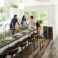 A modern kitchen with a large island, four stools, a family chatting at the counter, stainless steel appliances, and bright natural light.