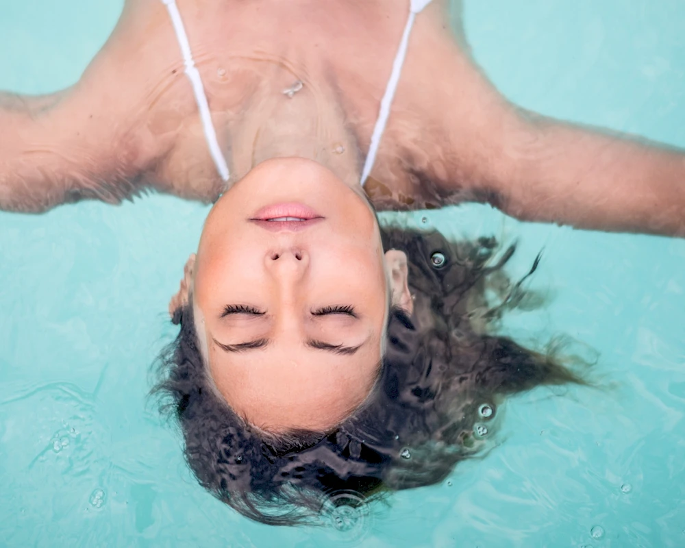 A person floating upside down in a turquoise pool, wearing a white swimsuit strap, with eyes closed and hair spread in the water.