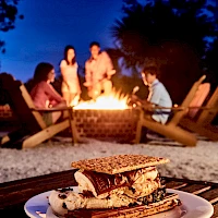 A cozy bonfire scene with friends around a fire pit at dusk, while a stack of grilled sandwiches sits on a plate in the foreground.