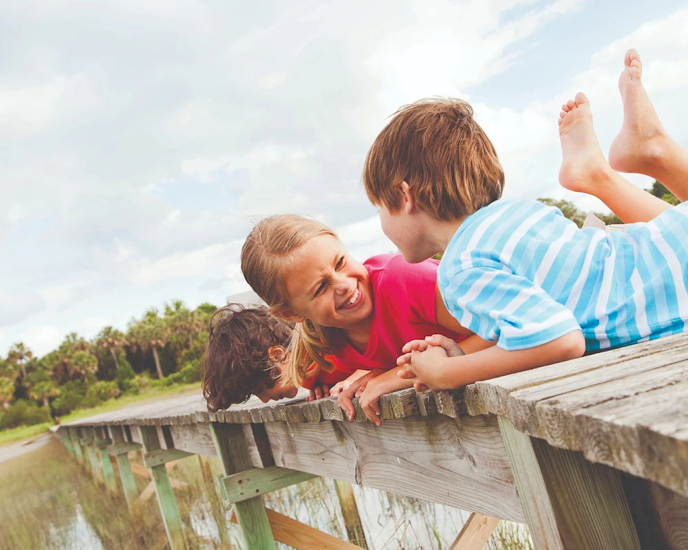 Three children are lying on a wooden railing beside a body of water, smiling and looking at each other.