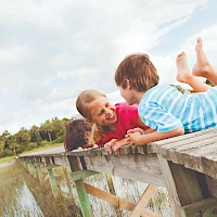 Three children are lying on a wooden railing beside a body of water, smiling and looking at each other.