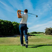 A golfer in mid-swing on a sunny golf course, blue sky, green fairway, and trees lining the course.