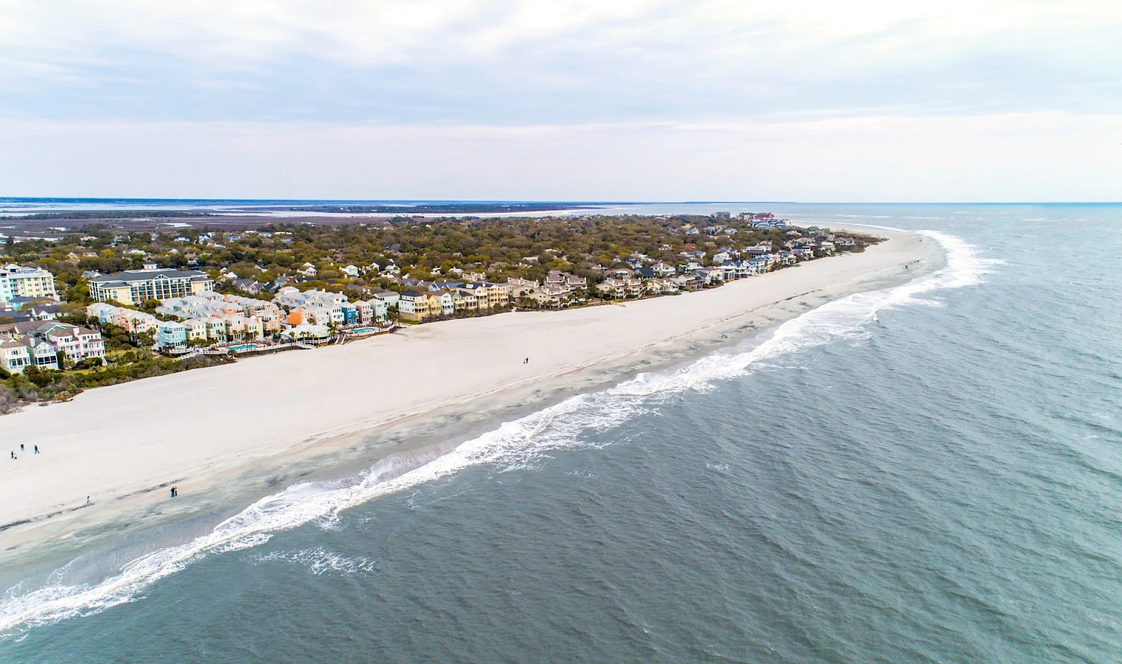 Coastline town with a sandy beach, row houses along the shore, and gentle waves curling onto the shore under a calm sky.