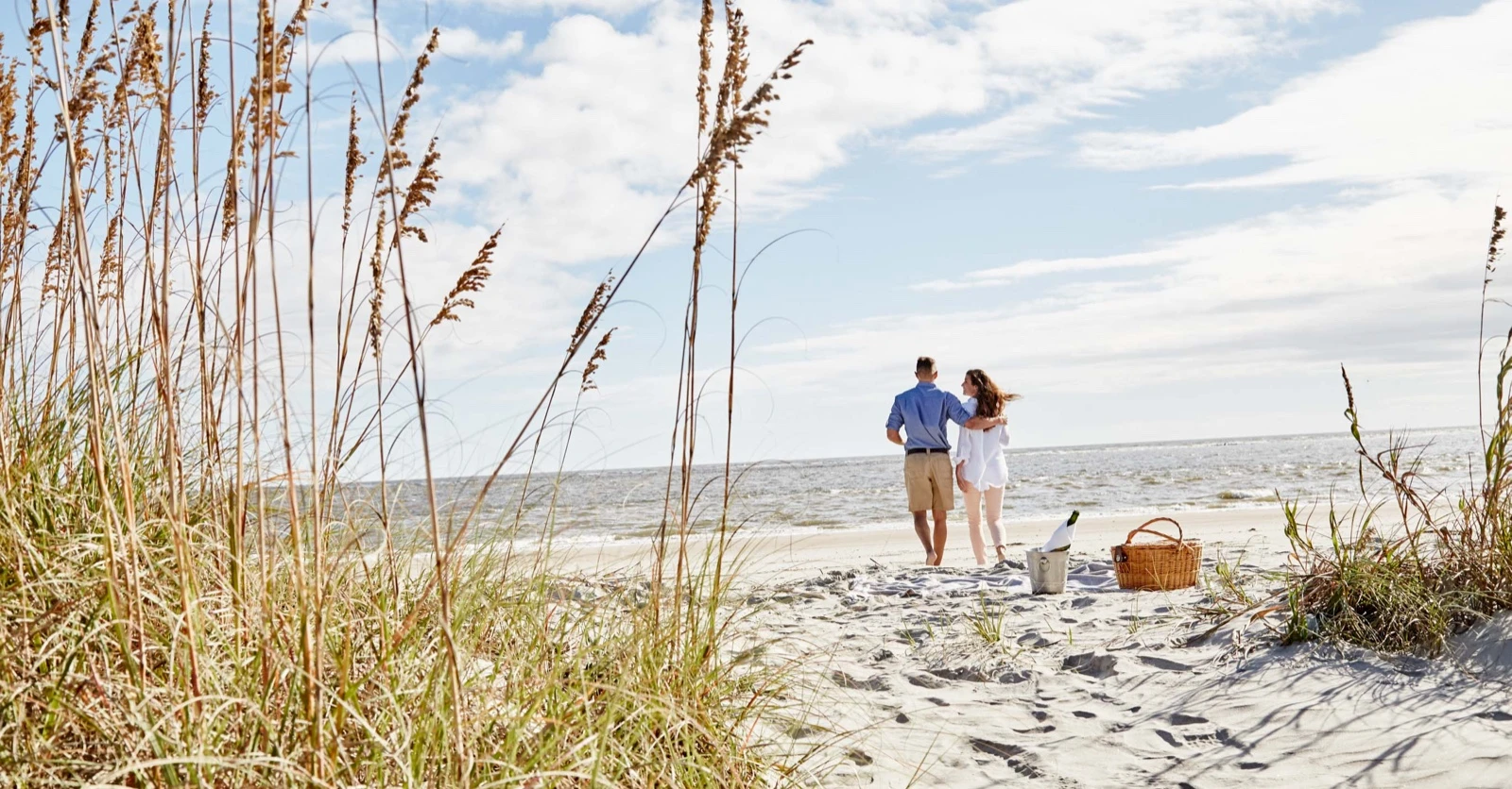 Two people walk arm-in-arm toward the sea on a sunny beach, with dune grasses in the foreground, a picnic basket nearby, and gentle waves.