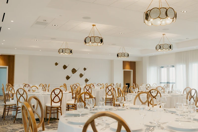 A bright banquet hall with round tables, white linens, and wooden chairs, set for a formal event with soft lighting.