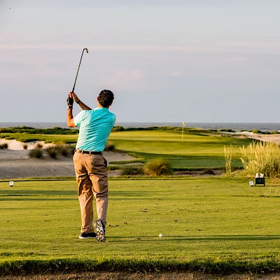 A golfer in a turquoise shirt and khaki pants swings a club on a green fairway, with dunes and shrubbery in the background.