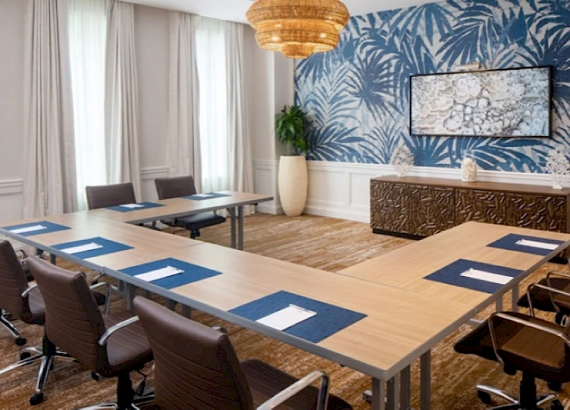 A modern conference room with a U-shaped table, blue place mats, white napkins, brown chairs, a bold blue botanical wallpaper, a large hanging light, and a decorative cabinet along the wall.