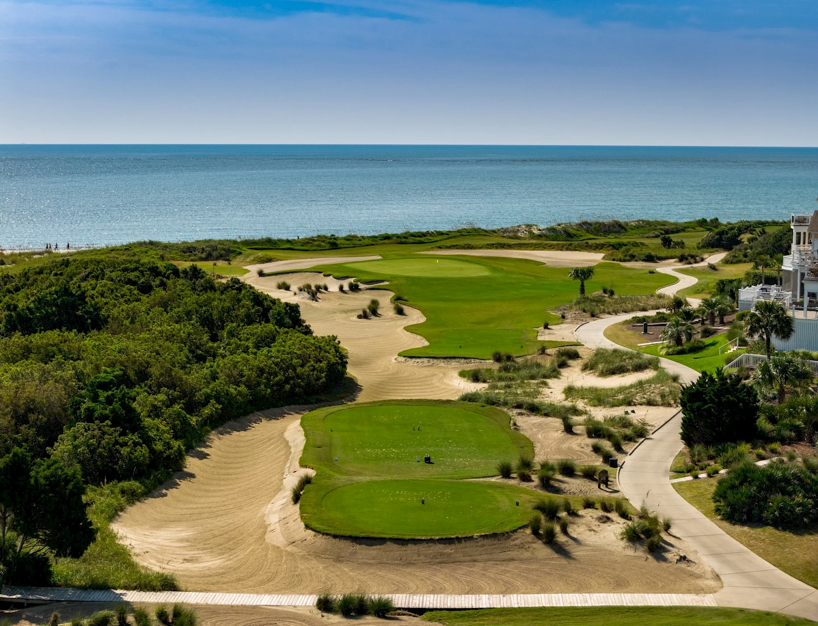 Coastal golf course with neatly trimmed greens, sandy bunkers, winding paths, lush shrubs, and the deep blue sea stretching to the horizon.