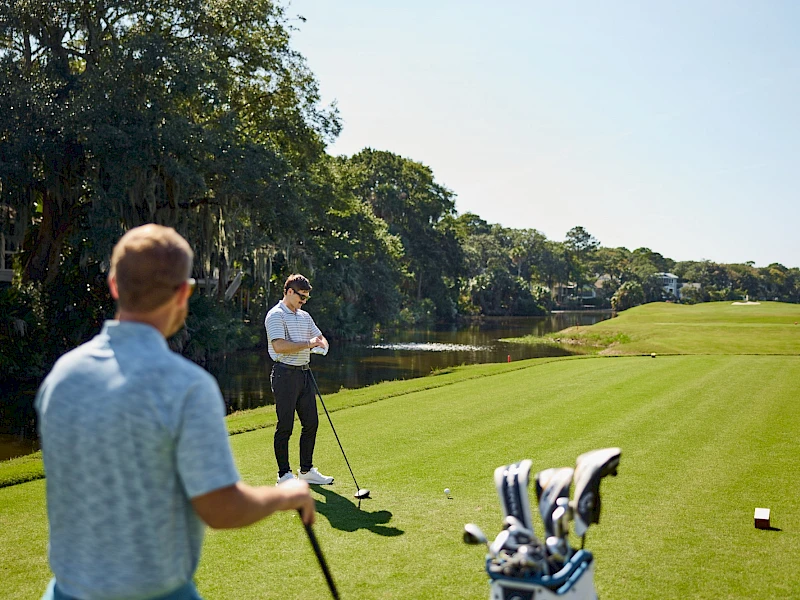 Two golfers practice on a sunny golf course: one man swings near a bag of clubs while another watches in the foreground, with greens and trees in the background.