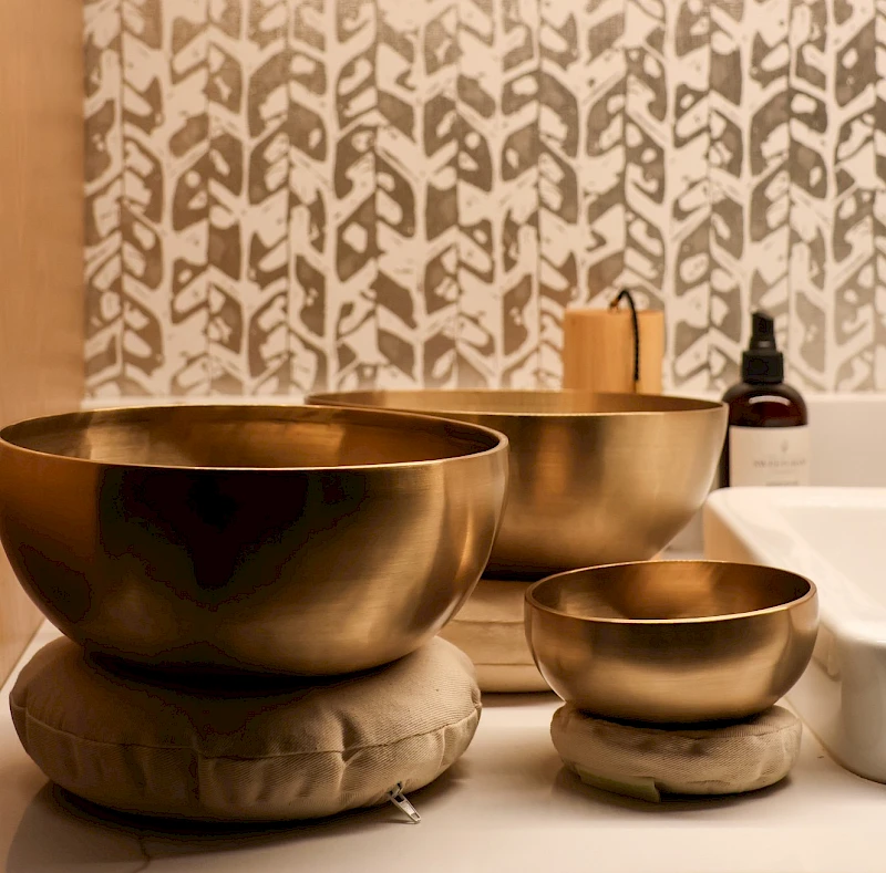 A serene spa-like scene with nested brass singing bowls, a small wooden mallet, and a bottle on a patterned wall backdrop.