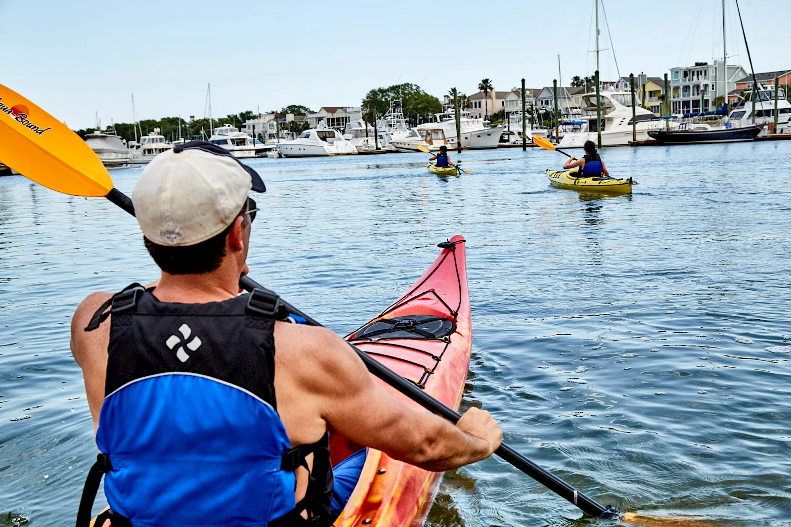 A person in a blue and black life vest kayaks in calm water, with others in small boats and a harbor with buildings in the background.