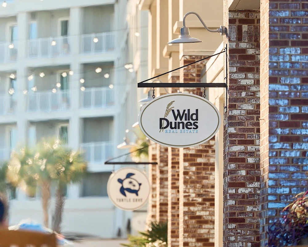 A street with brick building, hanging signs for Wild Dunes resort, outdoor lamps, and blurred people and balconies in the background.