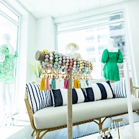 A bright boutique with a striped sofa, colorful beaded jewelry display, and green dress on a mannequin by a window, sunny and inviting.
