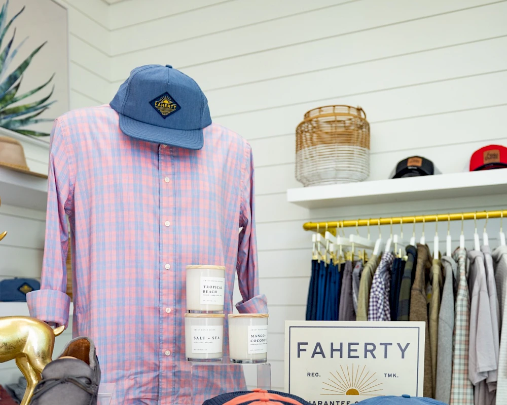 A pastel shop display with a pink plaid shirt on a mannequin, several colorful hats, folded clothes, and a sign reading "FAHERTY" in the background.