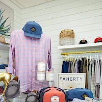 A pastel shop display with a pink plaid shirt on a mannequin, several colorful hats, folded clothes, and a sign reading "FAHERTY" in the background.