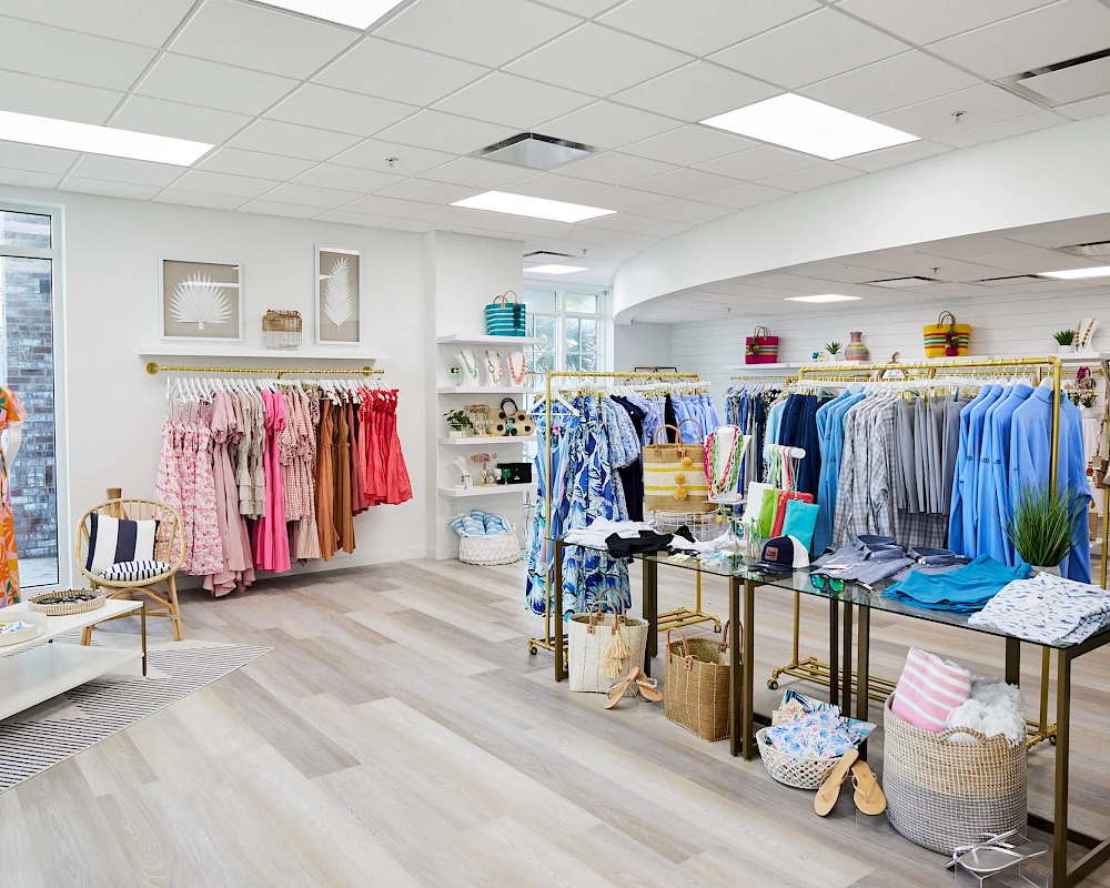 A bright boutique with light wood floors, racks of colorful dresses and tops, shelves of accessories, and a cozy seating area for shoppers.