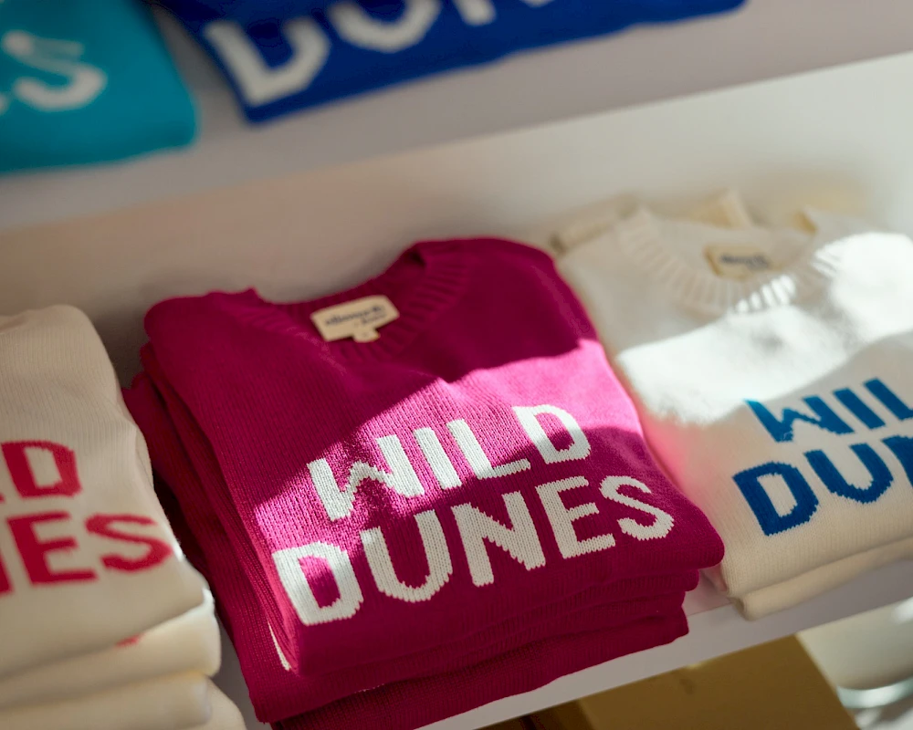 T-shirts on a rack with "WILD DUNES" in bold letters, white, pink, and cream colors, shop display at a clothing store.
