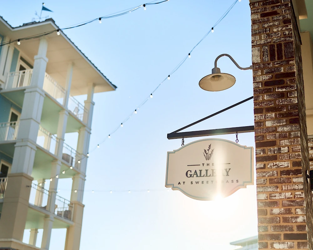 A sunny street scene featuring a brick building with a hanging lamp, string lights, and a sign that reads "Gallery" between pastel-colored apartment blocks.
