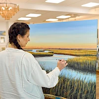 A woman in a light jacket paints a large landscape on a canvas in a bright gallery, featuring a sunset over marshland with tall grasses.