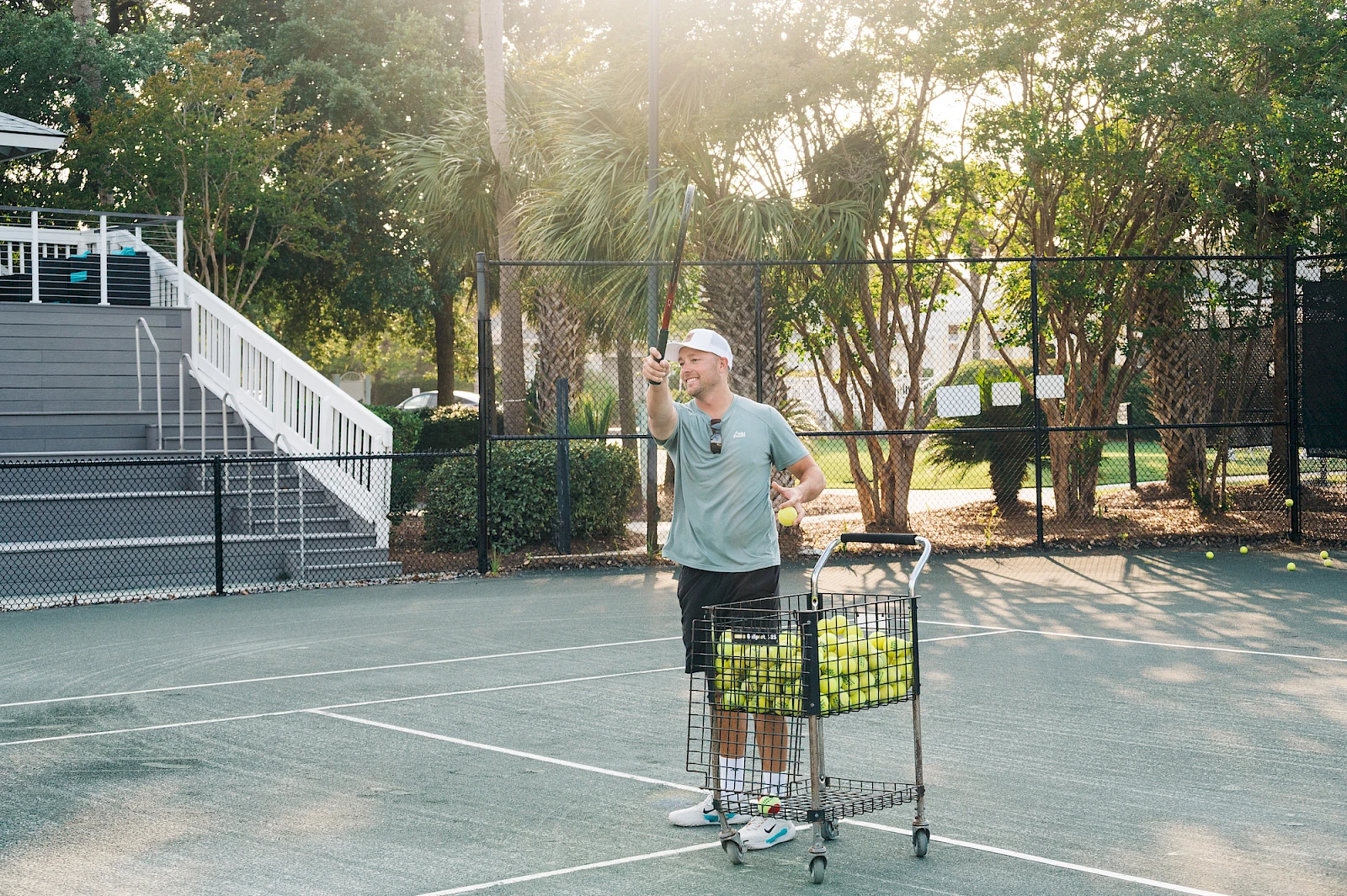 A man with a tennis cart stands on a court, raising one arm, wearing a cap and gray shirt, sunny park-like backdrop behind.
