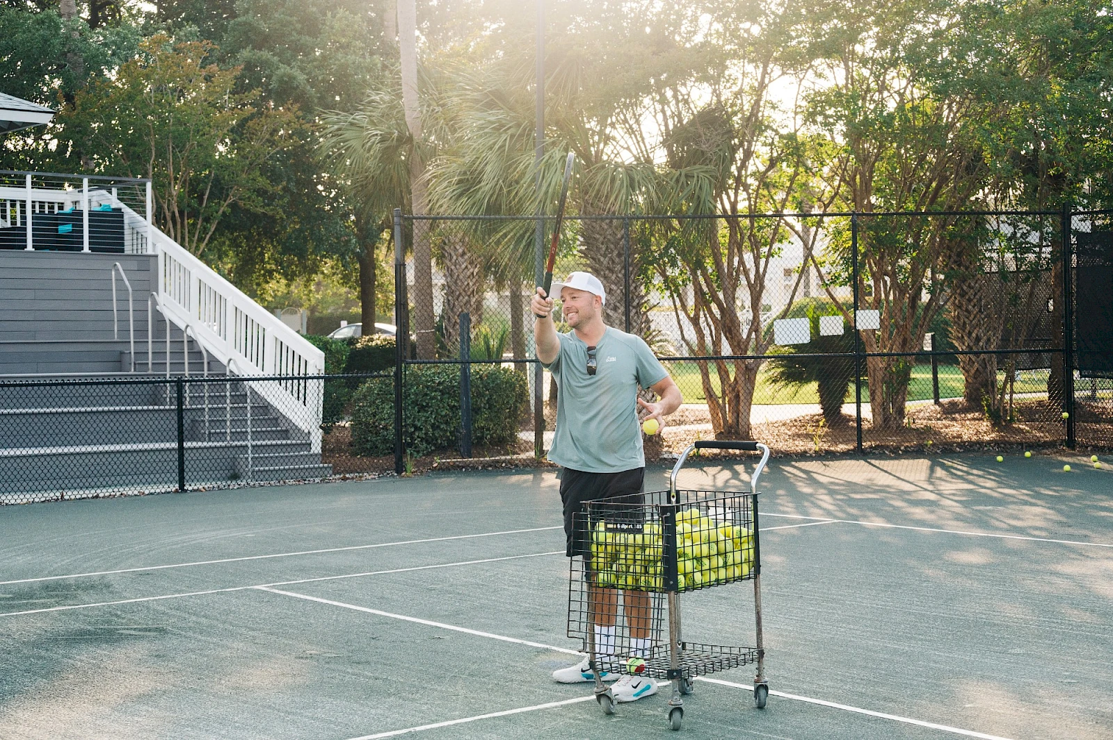 A man wearing a Santa hat stands on a tennis court with a cart full of yellow-green tennis balls, holding a ball in one hand and waving.
