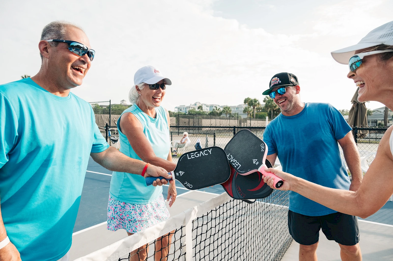 A group of four friends on a tennis court laughing and clinking paddles after a friendly game, wearing bright sporty outfits.