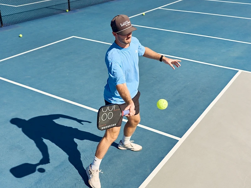 A man in a light blue shirt and cap is playing tennis on a blue court, preparing to hit a ball with a small clipboard in hand, in an outdoor setting.