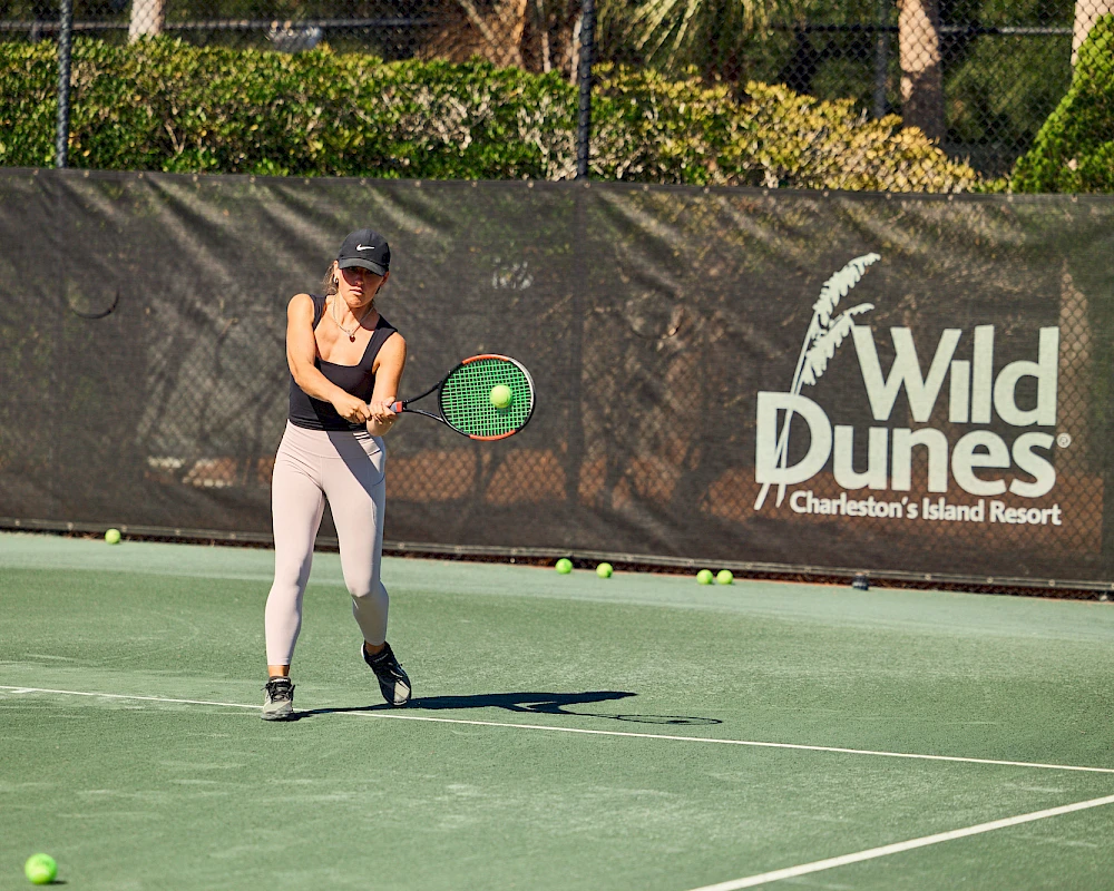 A female tennis player is hitting a backhand on an outdoor court, with a Wild Dunes banner in the background.