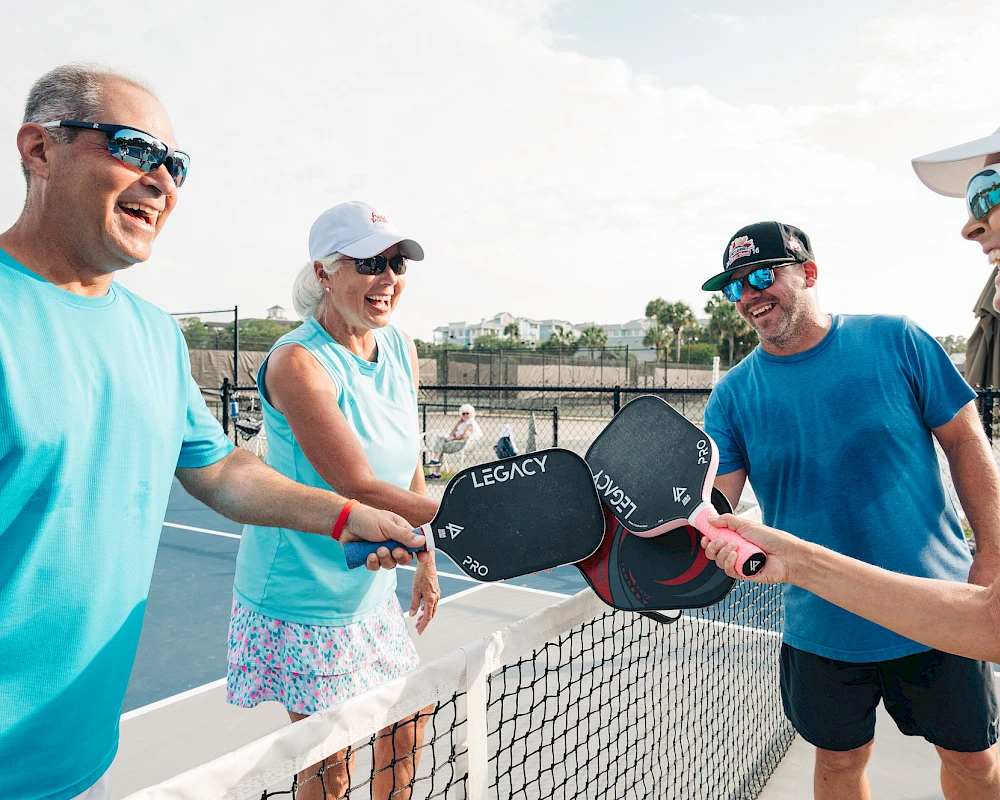 A group of four adults on a tennis court sharing smiles and high energy as they pass paddles and celebrate a game ending.
