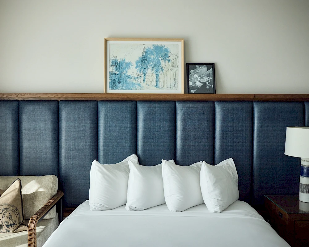 A neatly made bed with white pillows against a dark cushioned headboard, side table, lamp, and framed art above.
