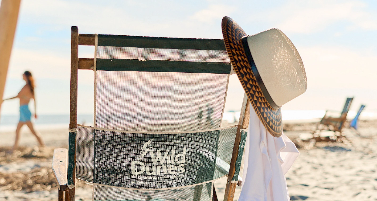 A sunlit beach scene with a worn chair facing the water, a wide-brimmed hat draped over it, and a person strolling along the shore.