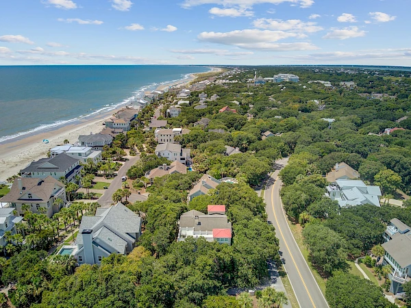 Aerial view of a coastal neighborhood with houses along a beach, lush green trees, winding roads, and the ocean on the left.