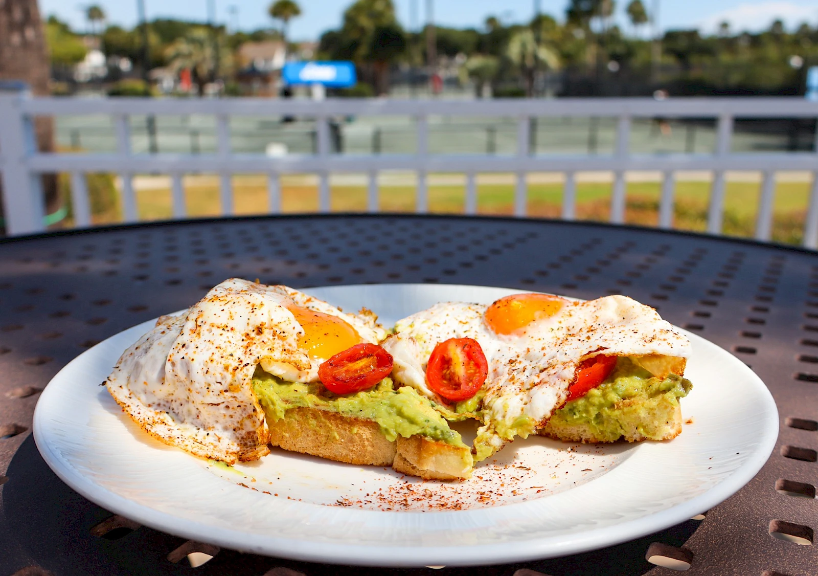 Two open-faced avocado toast slices topped with poached eggs and sliced chili on a white plate, lightly seasoned, outdoors, sunny day.