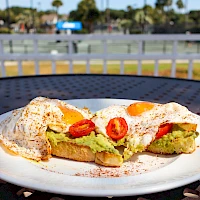 Two open-faced avocado toast slices topped with poached eggs and sliced chili on a white plate, lightly seasoned, outdoors, sunny day.