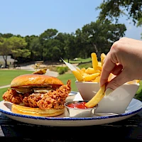A crispy fried chicken sandwich with fries and dipping sauces on a plate, outdoors on a sunny day.