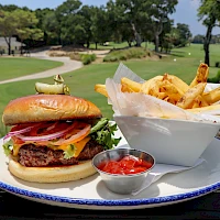 A juicy burger with bacon and tomato, crisp fries in a white pouch, and a small cup of ketchup on a blue-rimmed plate, outdoors.