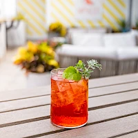 A bright red cocktail with ice and a mint sprig sits on a wooden table, blurred outdoor lounge in the background.
