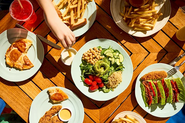 A colorful outdoor meal with fried chicken, salad, avocado toast, fries, and drinks on a wooden table.