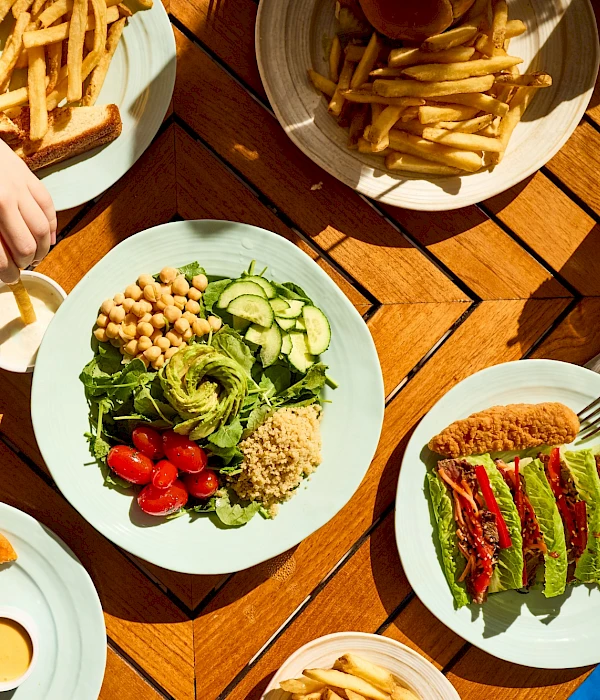 A colorful outdoor meal with fried chicken, salad, avocado toast, fries, and drinks on a wooden table.