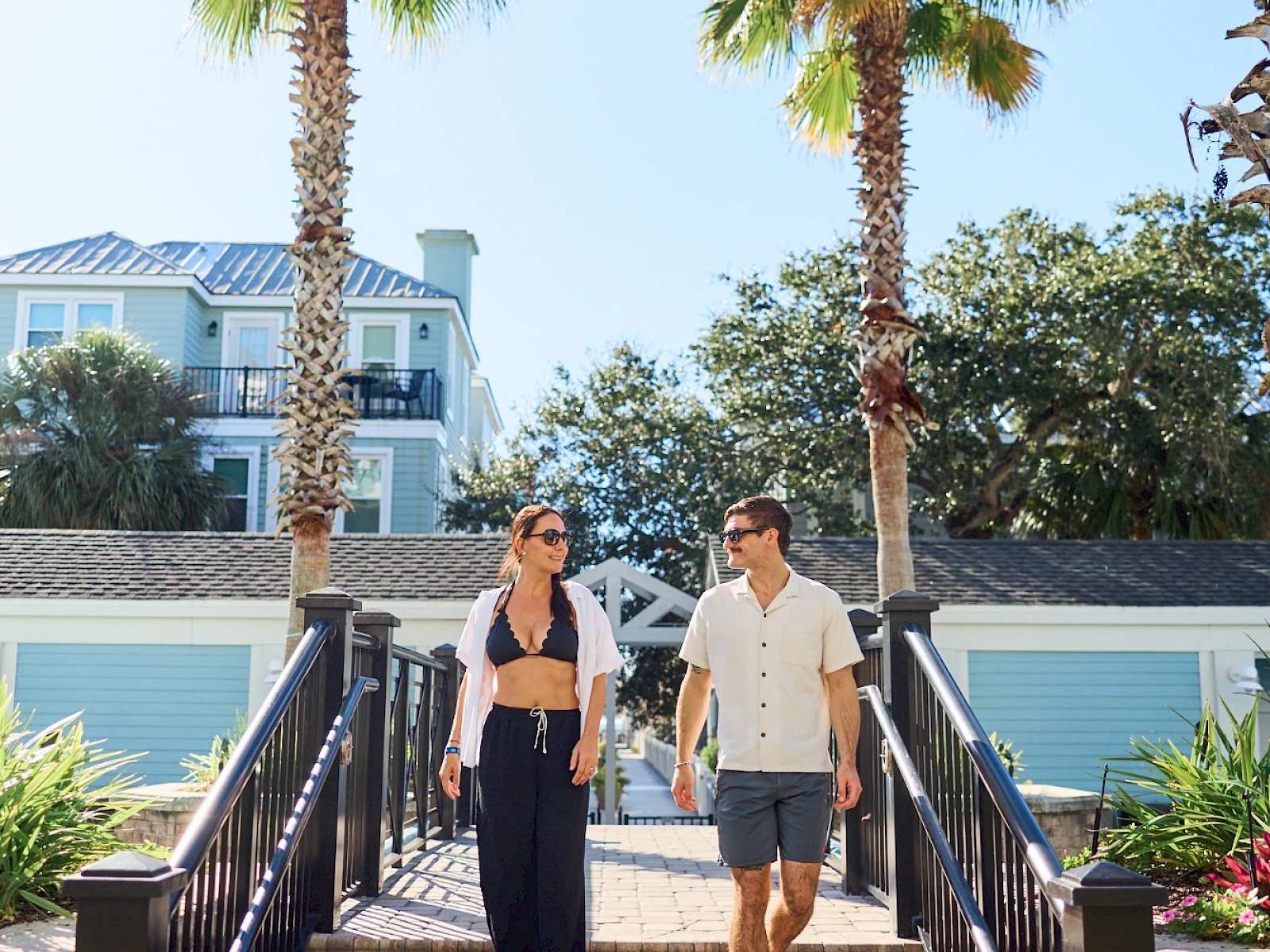 Two people walk down outdoor stairs between palm trees toward the camera, a sunny day with a bright blue sky and beachy homes in view.