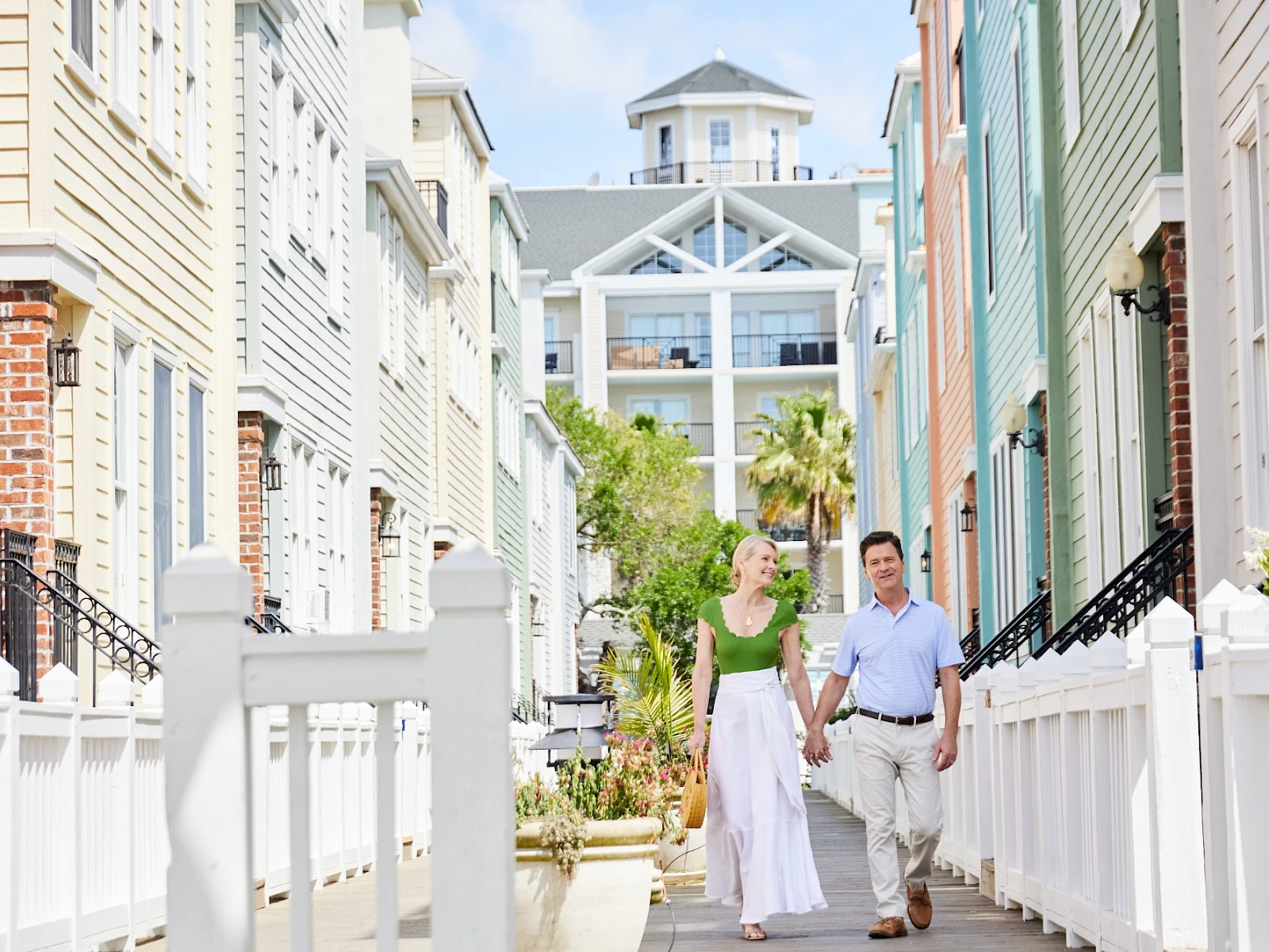 Two people walk hand-in-hand down a sunny, colorful alley lined with pastel houses and white fences. The scene feels like a charming coastal neighborhood.