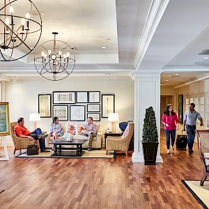A welcoming hotel lobby with a seating area, chandeliers, wooden floors, potted plants, and guests chatting while staff assist at the reception.