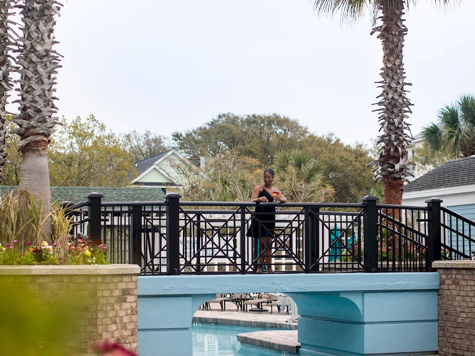 A resort pool with turquoise water, a decorative bridge, palm trees, blooming flowers, and a person standing on the bridge overlooking the pool.