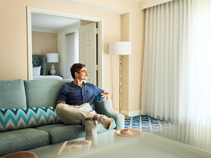 A man sits on a teal sofa in a bright hotel room, looking toward the window with sheer curtains, a lamp, and a mirror behind him.