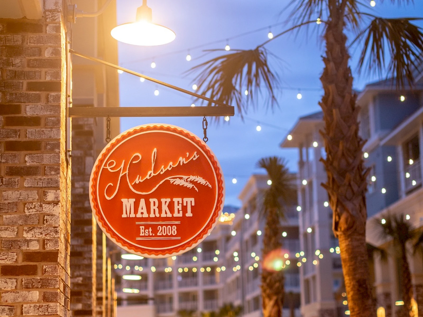 A red circular sign for a market hangs by a brick storefront at dusk, palm trees, string lights, and a sidewalk cafe vibe.