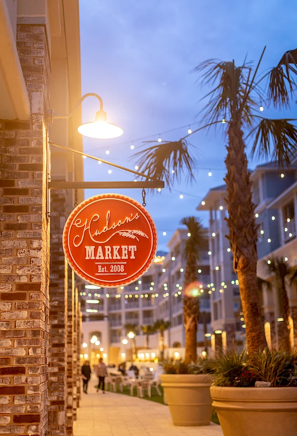 A brick-sided storefront with a round red sign reading “Gadsden Market Est. 2002” hangs outside, string lights, palm trees, and warm evening ambience.
