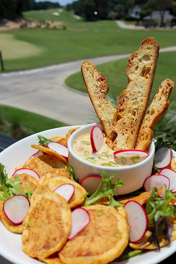 A platter of crispy potato chips and radishes with a creamy dip, garnished with greens, alongside toasted baguette slices.
