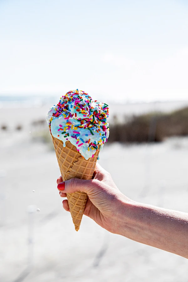 A hand holds a colorful ice cream cone with blue icing and rainbow sprinkles, at a beach.